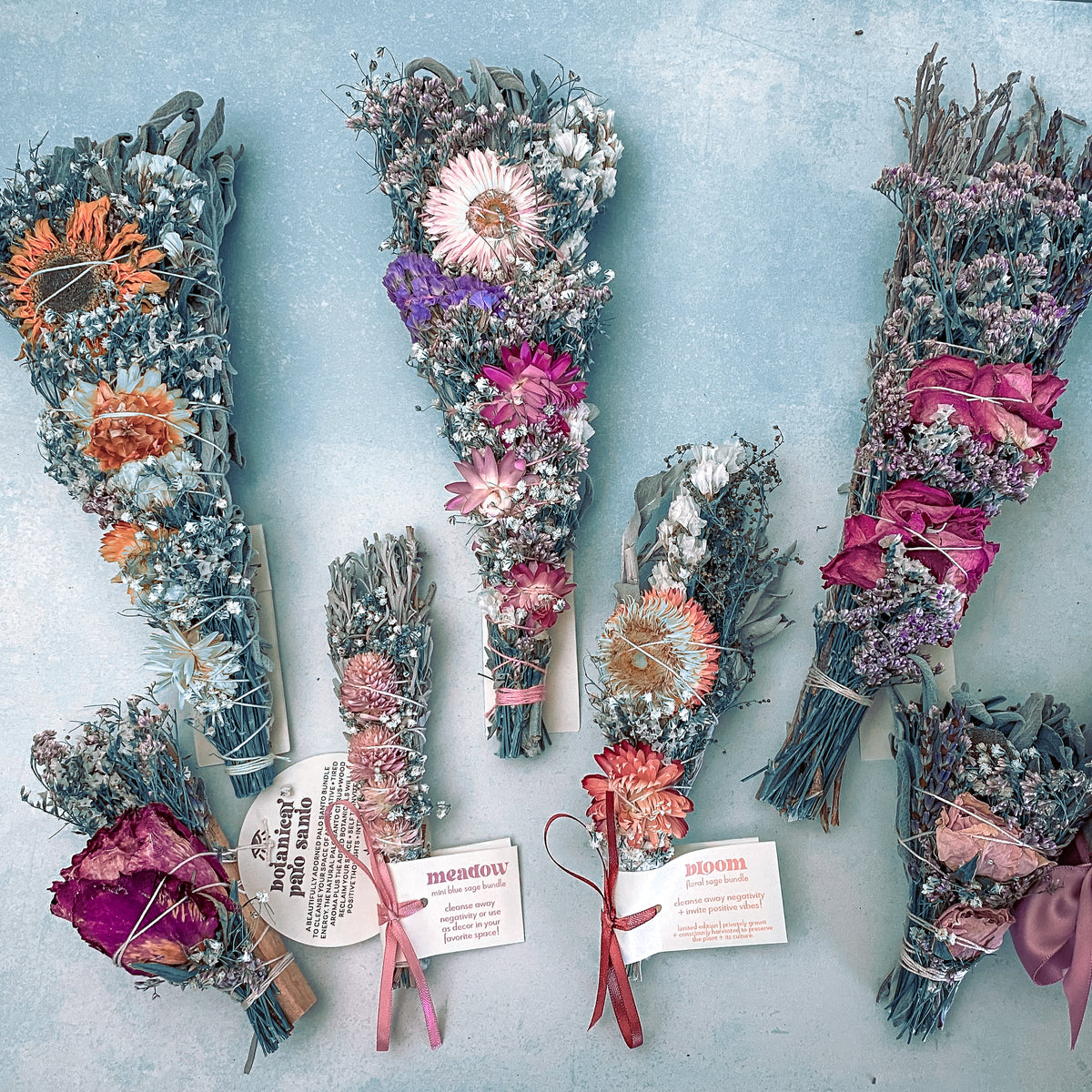 Six bundles of dried flowers in various sizes, featuring bright pink, purple, and yellow blooms, are arranged on a light blue surface. Two bundles have labels reading meadow and bloom.