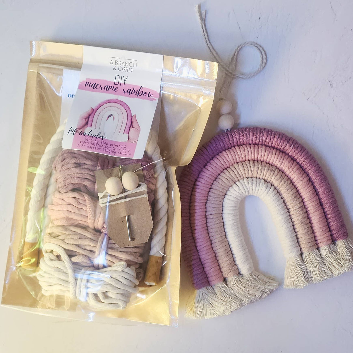 The Sweet Pink DIY Rainbow Beginner Macrame Kit by A Branch & Cord comes in a clear bag and includes everything to create a pink, cream, and purple wall hanging rainbow with tan string and wooden beads for easy display.