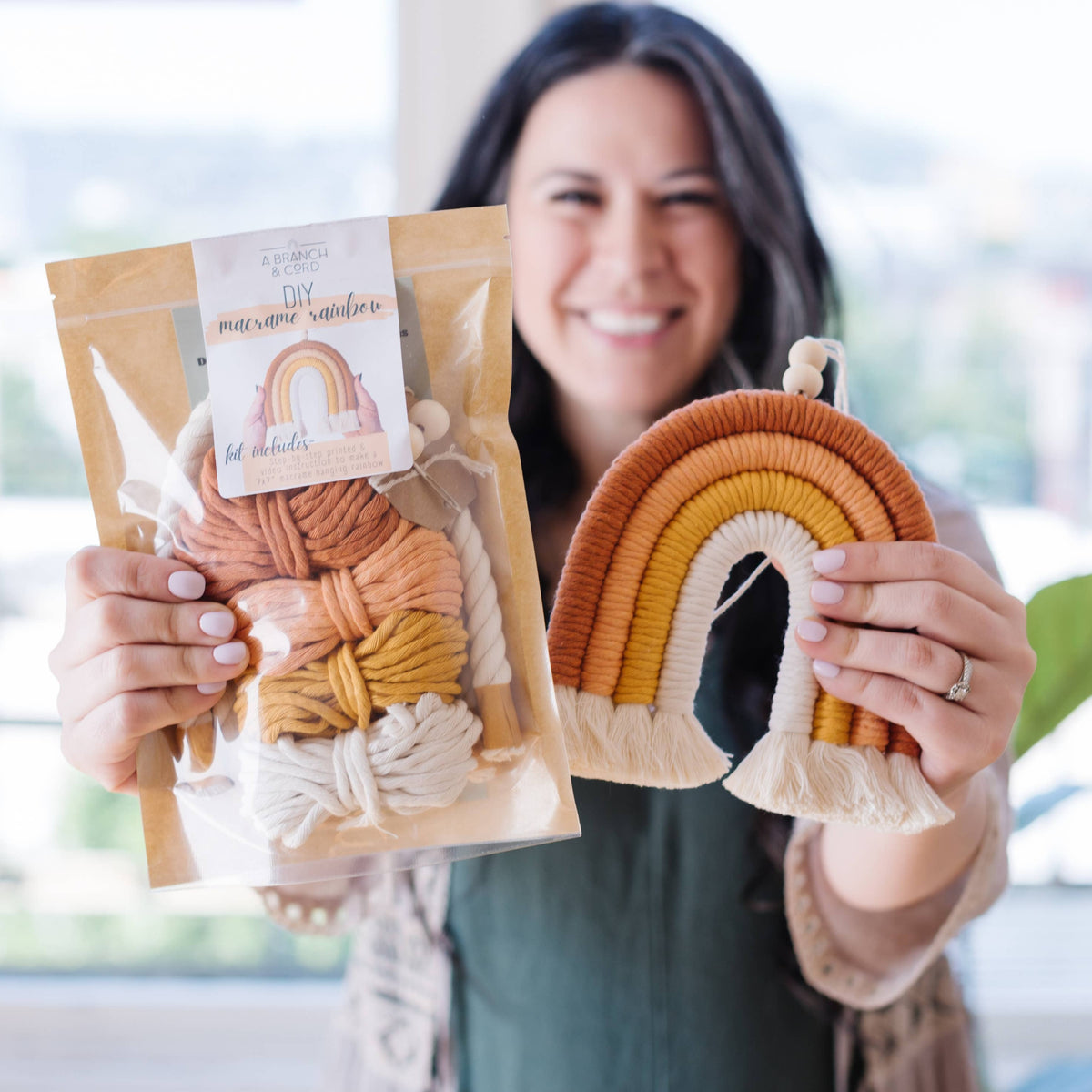 A smiling woman holds the Sweet Pink DIY Rainbow Beginner Macrame Kit by A Branch & Cord in one hand and a finished earthy-toned rainbow wall hanging in the other, highlighting her crafting skills in a bright indoor space.