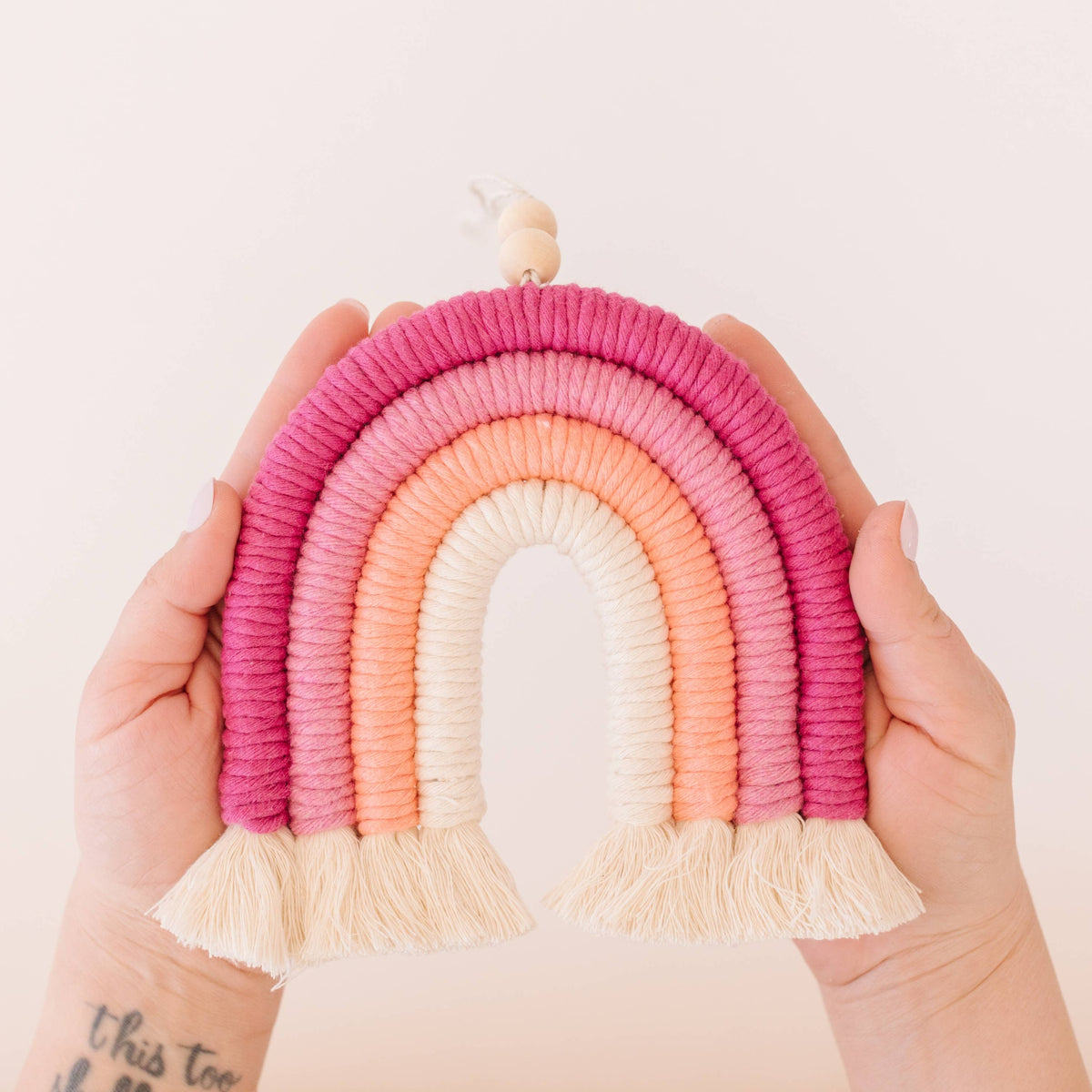 Two hands display the Sweet Pink DIY Rainbow Beginner Macrame Kit by A Branch & Cord—a handmade woven wall hanging in pink, orange, and cream with tasseled ends, capturing the joy of creating your own rainbow decor. Light background. Tattoo on wrist visible.