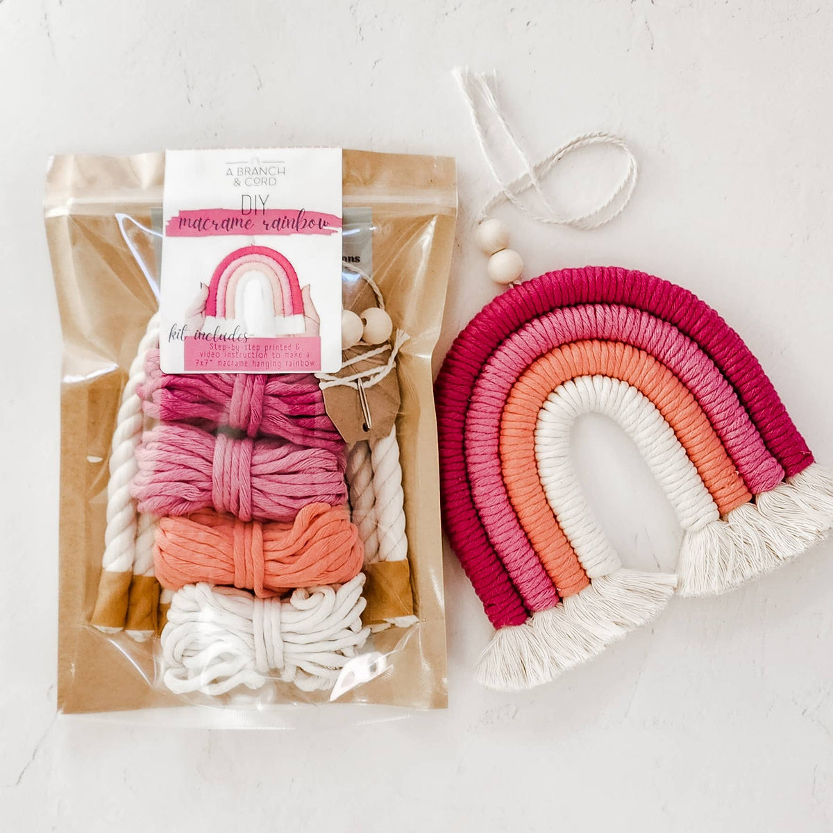 The Very Berry DIY Rainbow Beginner Macrame Kit by A Branch & Cord comes in a clear package with pink, orange, and white cords, shown next to a finished rainbow wall hanging with matching colors and beaded string—perfect for beginner crafters.
