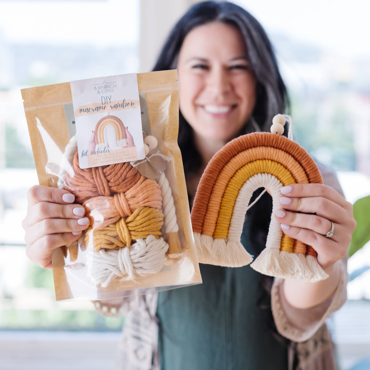 A smiling woman holds the Very Berry DIY Rainbow Beginner Macrame Kit by A Branch & Cord in one hand and a completed wall hanging in earthy orange, brown, and cream tones in the other, showcasing a delightful crafting experience.
