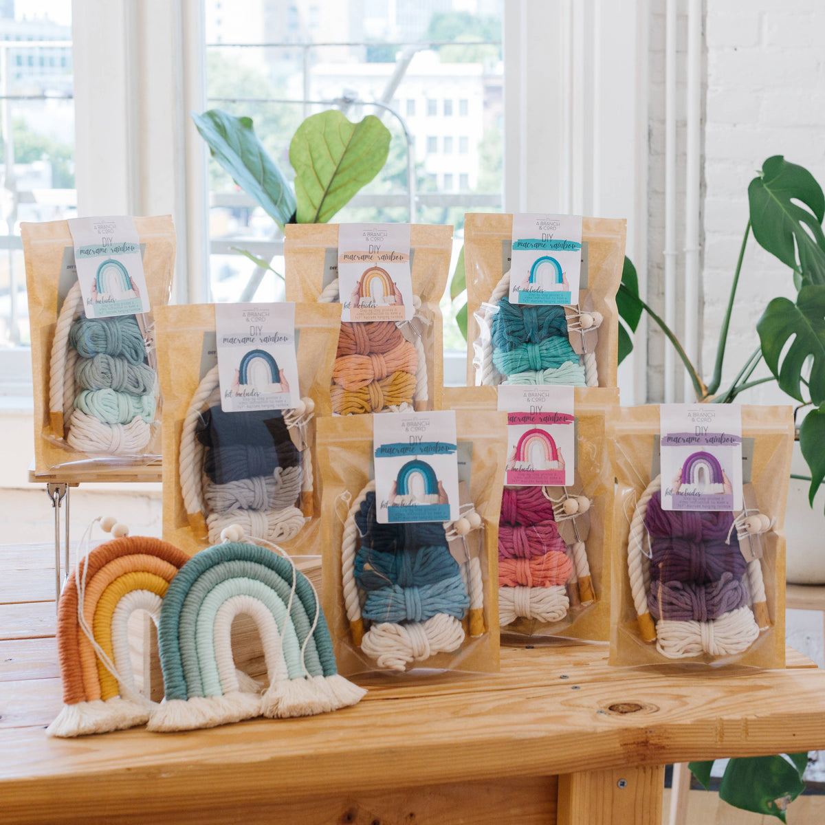 Several A Branch & Cord Very Berry DIY Rainbow Beginner Macrame Kits in vibrant colors are displayed on a wooden table beside finished rainbow wall hangings, with natural light and greenery enhancing the inviting crafting space.