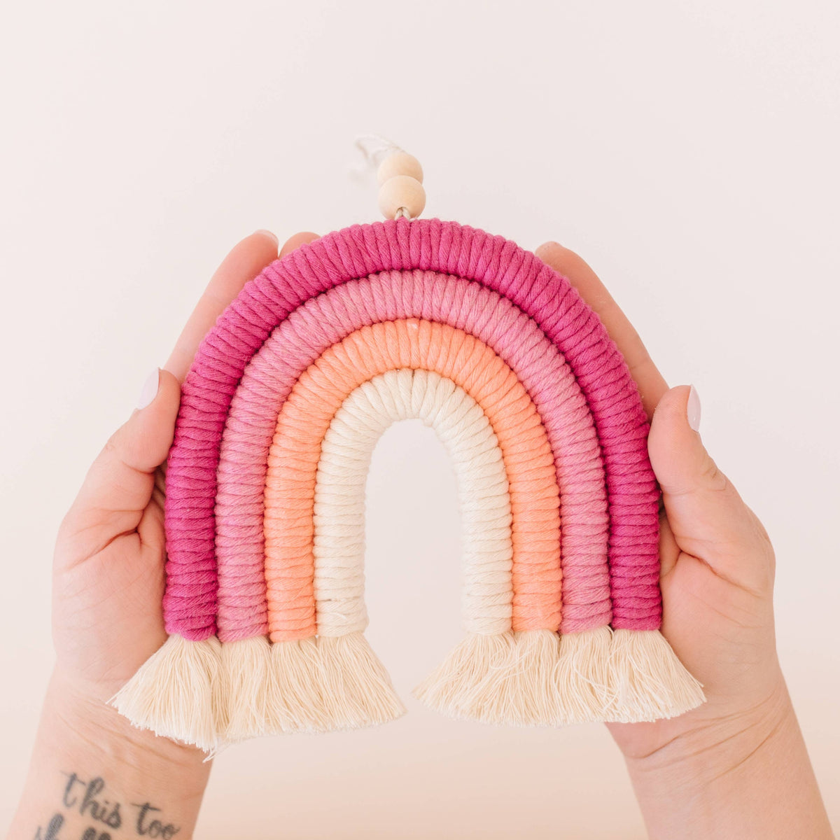 Two hands hold the Very Berry DIY Rainbow Beginner Macrame Kit by A Branch & Cord—a decorative, woven rainbow in pink, peach, and cream with tassels. Part of a tattoo is visible on one wrist against a soft, light background.