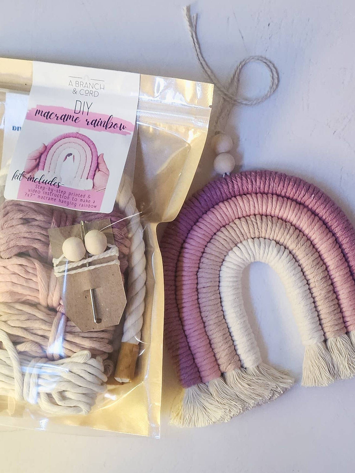 The Sweet Pink DIY Rainbow Beginner Macrame Kit by A Branch & Cord features pink, purple, and cream cords, wooden beads, and instructions beside a finished soft-tone rainbow wall hanging with fringe for a delightful crafting experience.