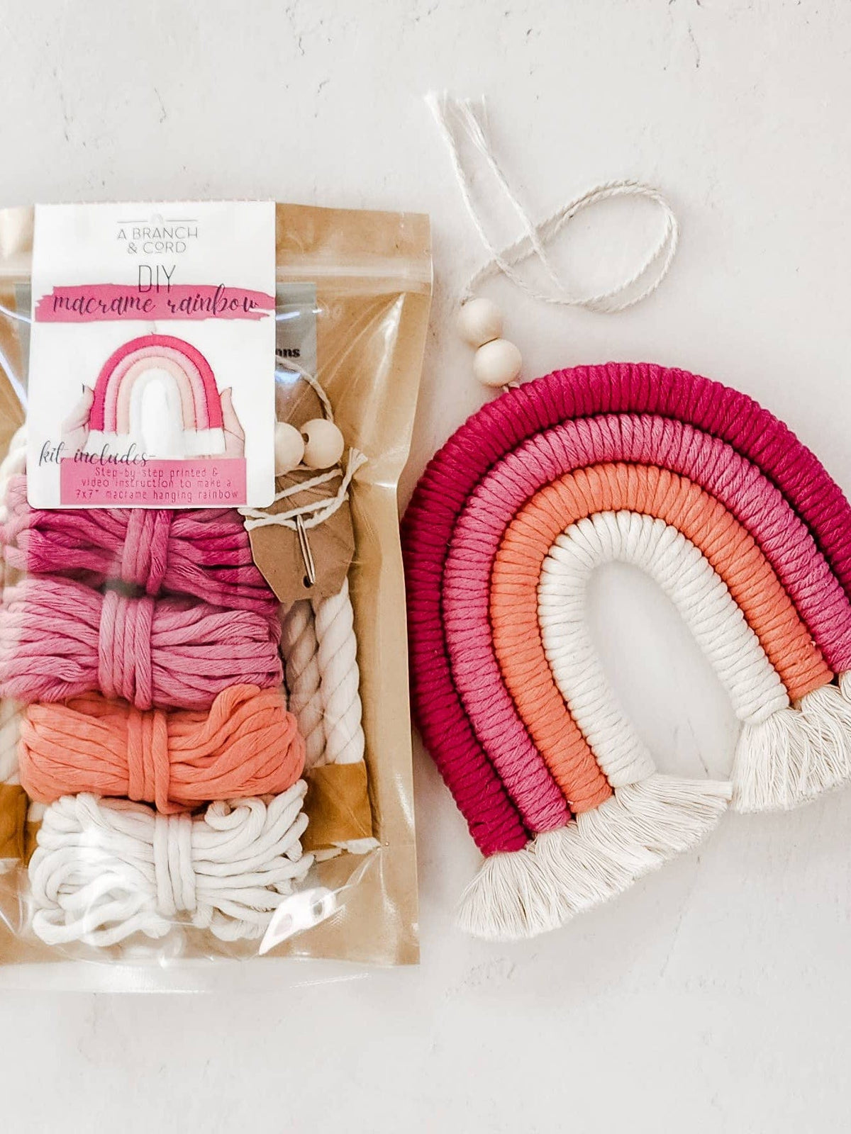 The A Branch & Cord Very Berry DIY Rainbow Beginner Macrame Kit in a clear package is shown next to a finished pink, peach, and white wall hanging with fringe and two wooden beads, perfect for charming craft projects.