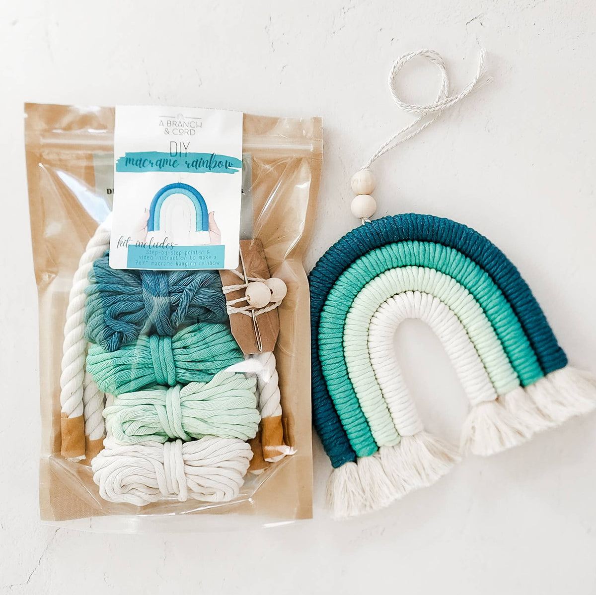 A Branch & Cord’s Teal DIY Rainbow Beginner Macrame Kit comes in a clear package with green, blue, and white yarn and cords. Beside it is a finished macrame rainbow decoration with matching colors and small wooden beads.