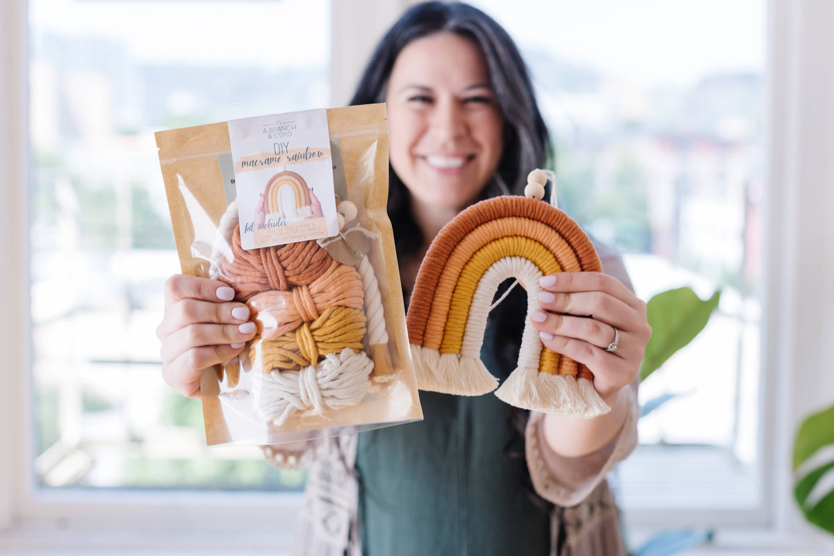 A smiling woman holds up the A Branch & Cord Teal DIY Rainbow Beginner Macrame Kit with yarn and a pattern, standing in a sunlit room ready for the perfect crafting experience.