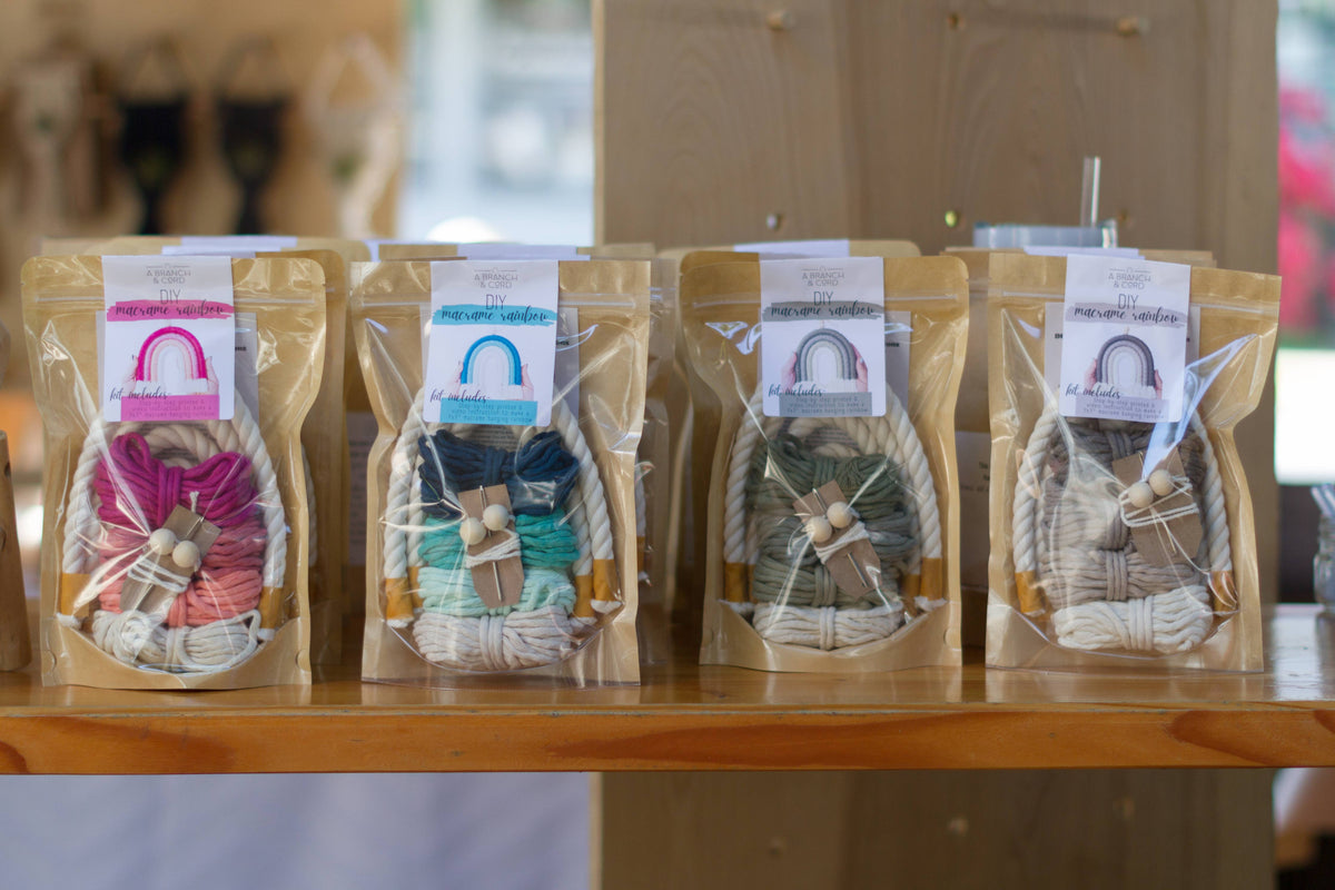 Four boxes of A Branch & Cord Teal DIY Rainbow Beginner Macrame Kits are displayed upright on a wooden shelf, each with a rainbow-themed tag, offering a colorful and fun macrame crafting experience.