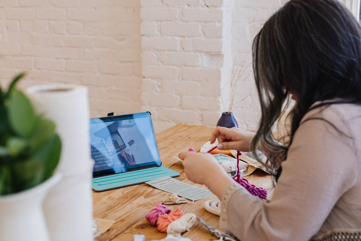A person with long dark hair sits at a wooden table, crafting with colorful yarn from the Teal DIY Rainbow Beginner Macrame Kit by A Branch & Cord, while watching a tutorial on a tablet amid supplies and plants in sunlit natural light.