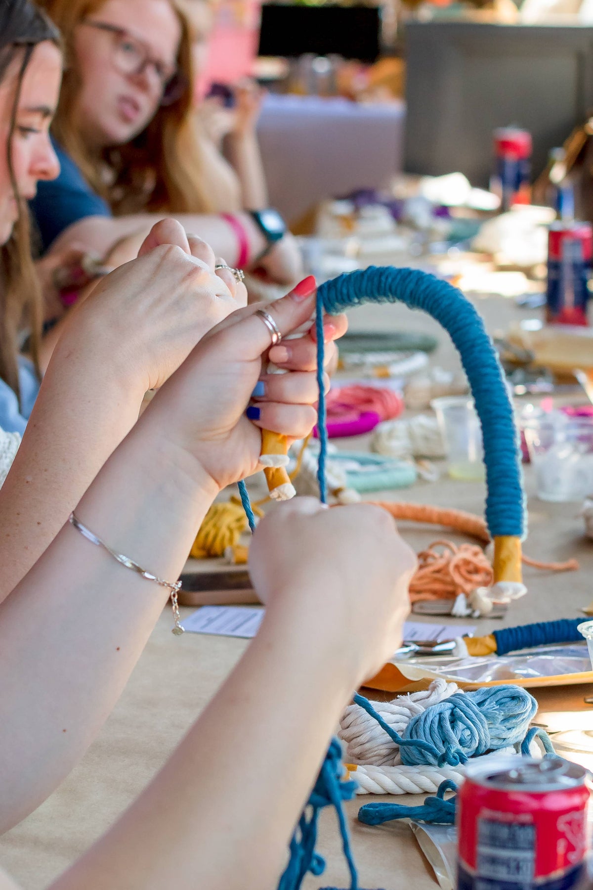 Several people sit around a table crafting with the Teal DIY Rainbow Beginner Macrame Kit by A Branch & Cord, wrapping blue yarn around a curved form with colorful yarns and kits scattered nearby. A can of soda sits in the foreground.