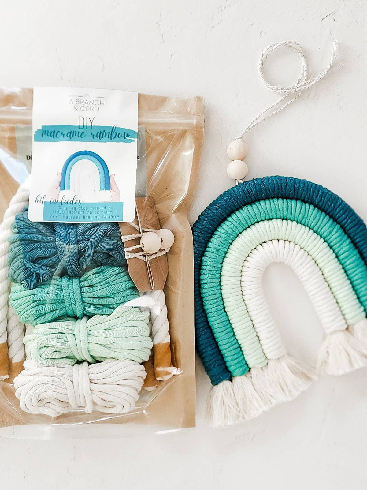 The Teal DIY Rainbow Beginner Macrame Kit by A Branch & Cord includes blue, green, and white cords, wooden beads, and a finished rainbow—all beautifully arranged on a light background for a fun crafting experience.