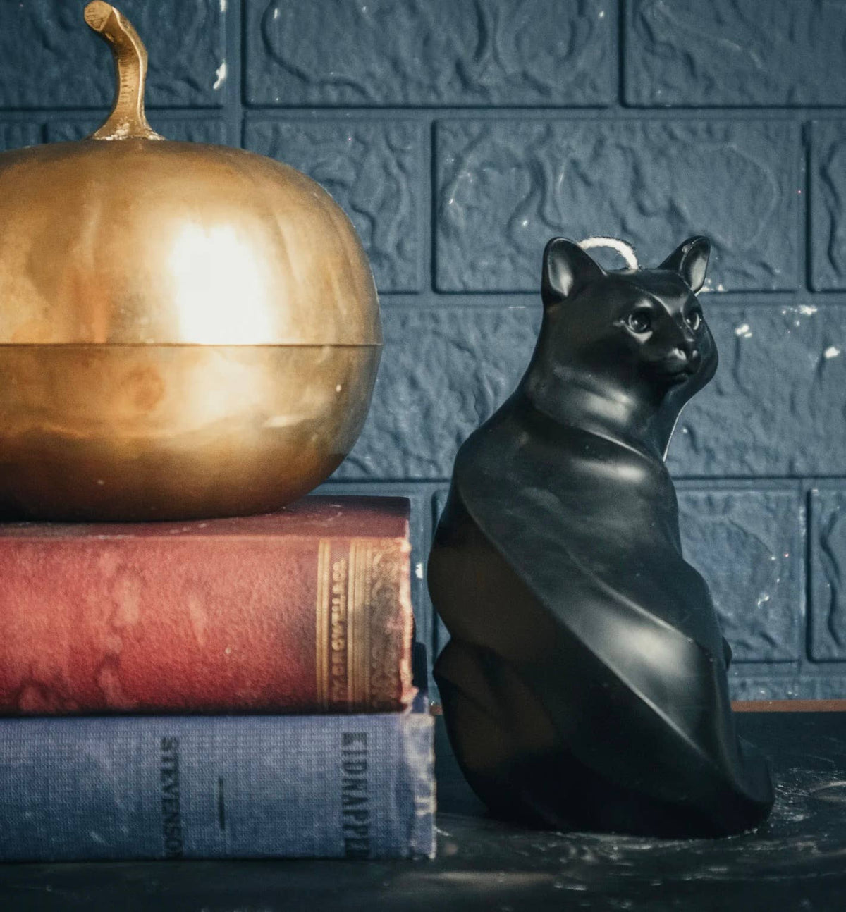 The A Pleasant Thought Black Cat Pillar Candle sits beside a golden apple-shaped container on two vintage books, set against a dark blue textured brick wall.