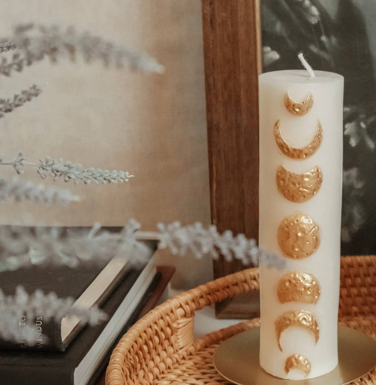 The Golden Moon Phase Pillar Candle by A Pleasant Thought, made from white soy wax and adorned with golden moon phases, rests on a woven tray beside stacked books, foliage in the foreground, and a framed picture behind.