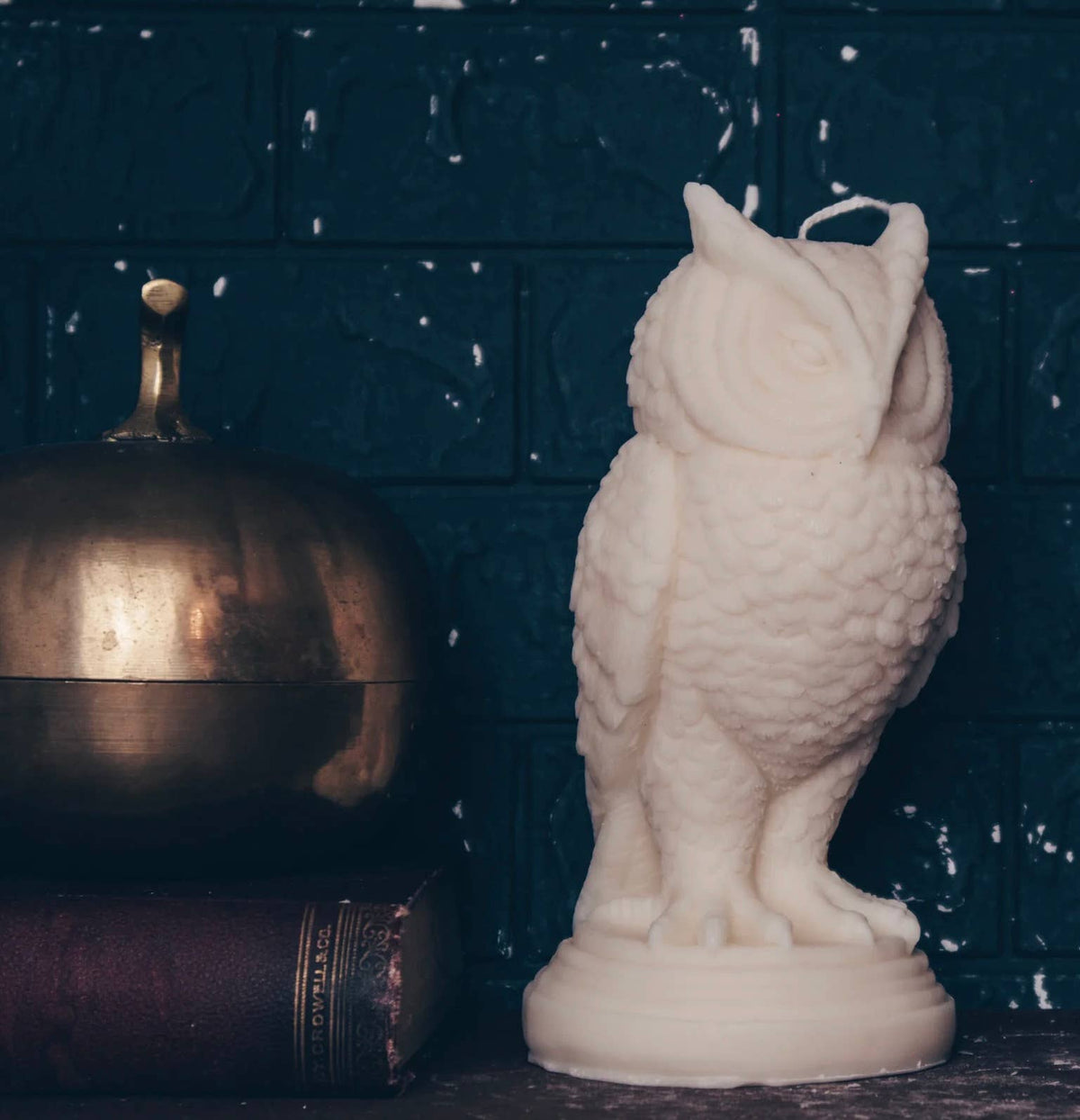The Snowy Owl Pillar Candle by A Pleasant Thought rests beside a closed brass container and a dark brown book, set against a textured dark blue brick wall.
