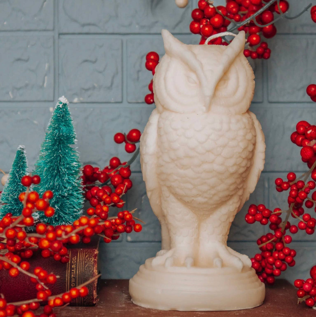 The Snowy Owl Pillar Candle by A Pleasant Thought, handcrafted in Canada from soy wax, sits on wood beside two mini pine trees, red berries, and a closed book, set against a blue textured wall.