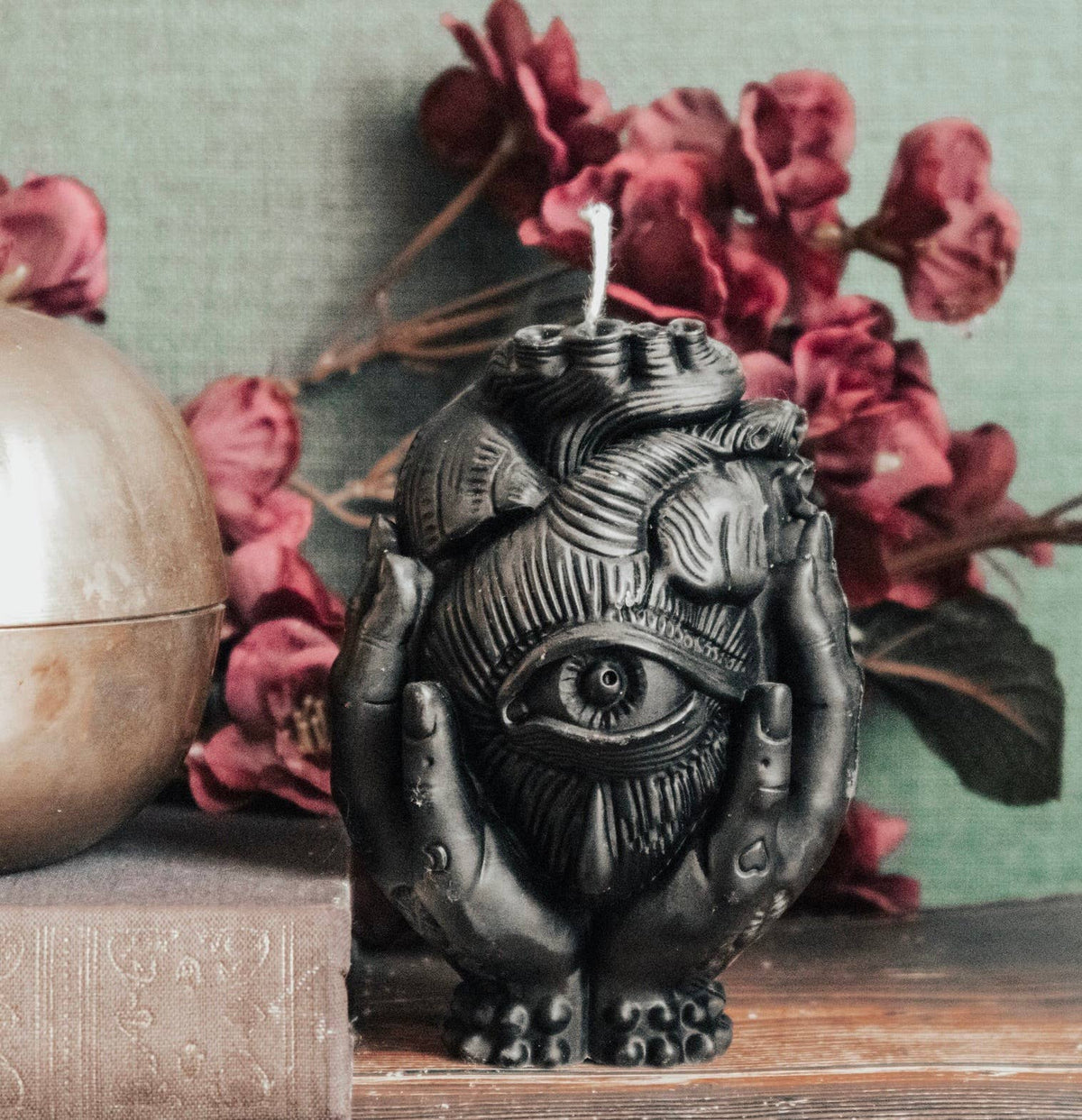 The Third Eye Heart Pillar Candle by A Pleasant Thought is a handcrafted soy wax candle shaped like a black heart with a central third eye, held by sculpted hands. Red flowers, a gold sphere, and a brown book rest on the wooden surface behind it.