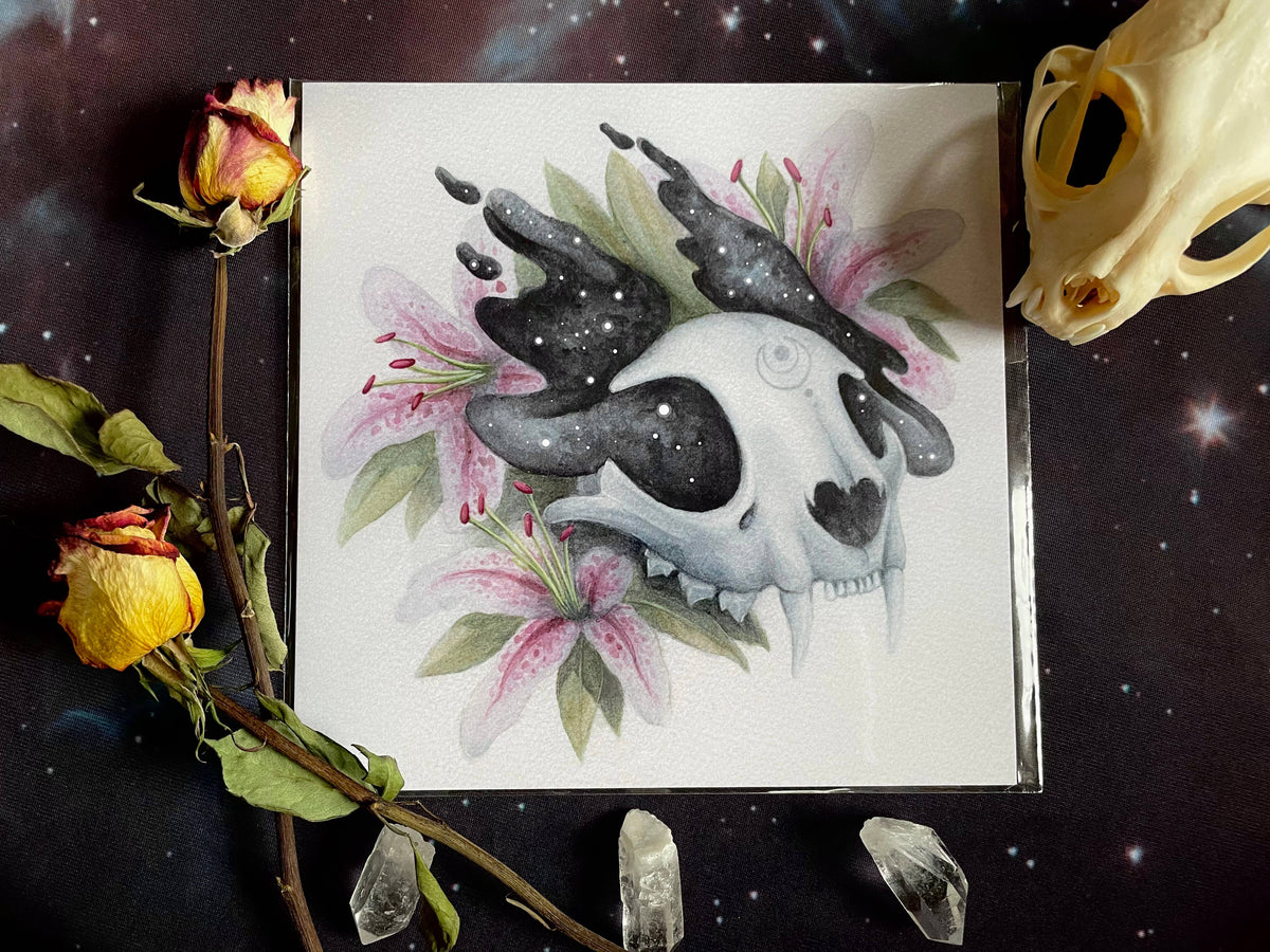 The Luna Art Print (8 x 8) by Amanda Tulacz Illustration features a stylized animal skull with starry black antlers and pink lilies, printed on archival cardstock and elegantly displayed with space-inspired decor.