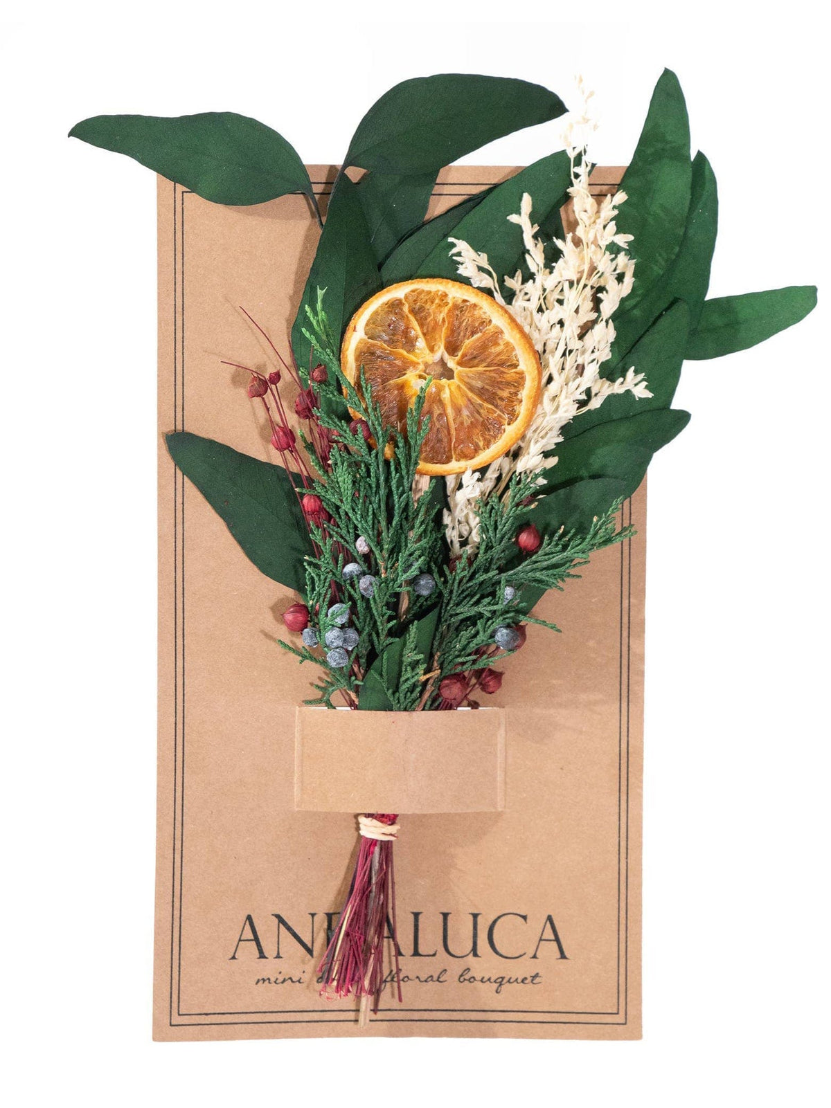 The Winter Citrus Mini Bouquet by Andaluca features green leaves, white and red sprigs, and a natural orange slice on a brown card—an ideal decorative accent for any space.