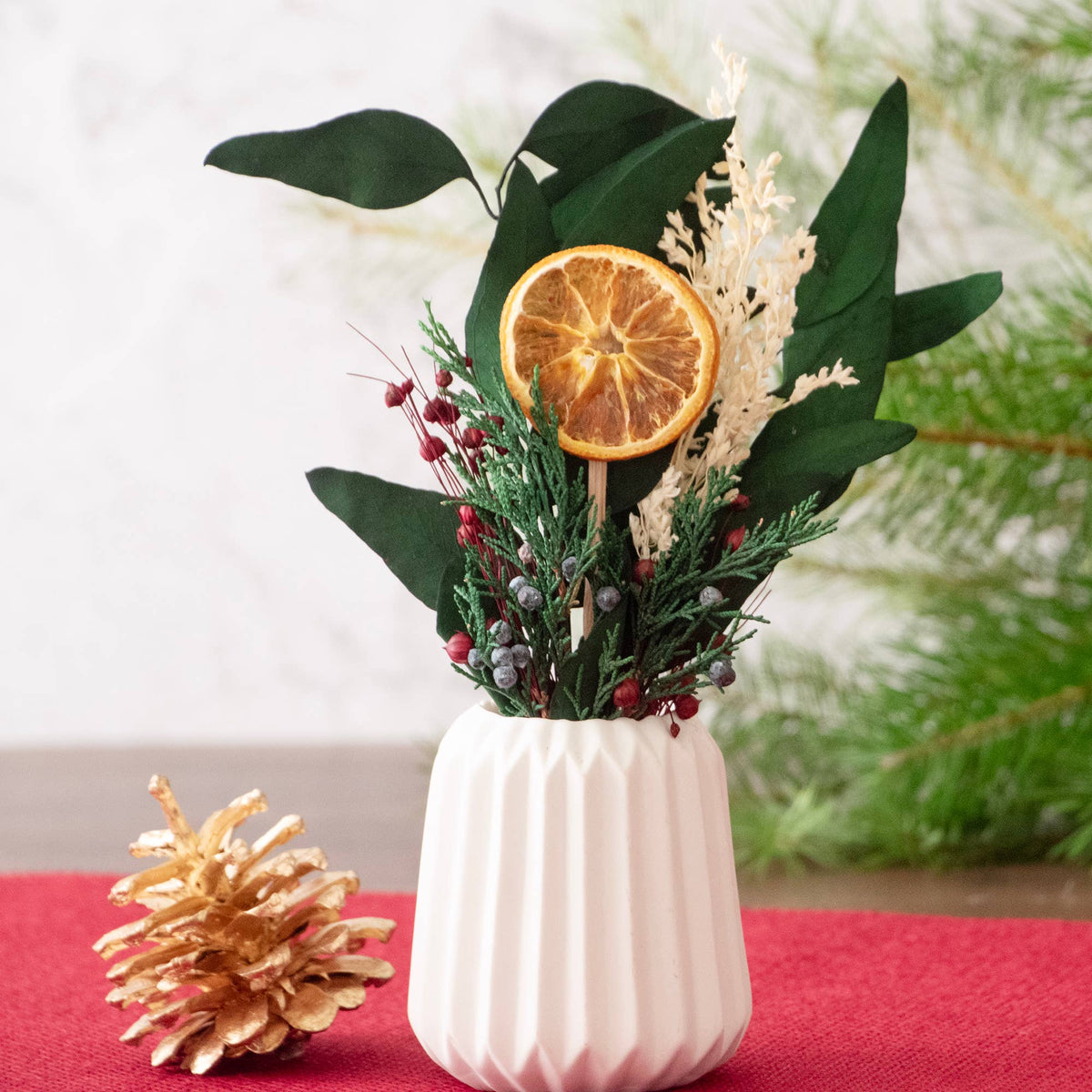 The Andaluca Winter Citrus Mini Bouquet sits in a small white vase, featuring artificial greenery, red berries, white dried flowers, and a natural orange slice. A gold-painted pine cone accents the red placemat while blurred pine branches appear in the background.