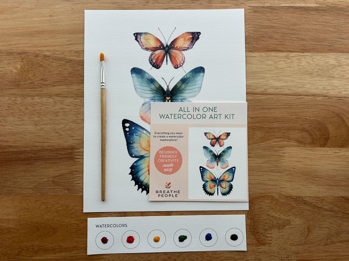 The Breathe People Butterflies All-In-One Beginner Watercolor Kit includes a butterfly design sheet, paintbrush, booklet, and six-color watercolor strip, all displayed on a wooden table.