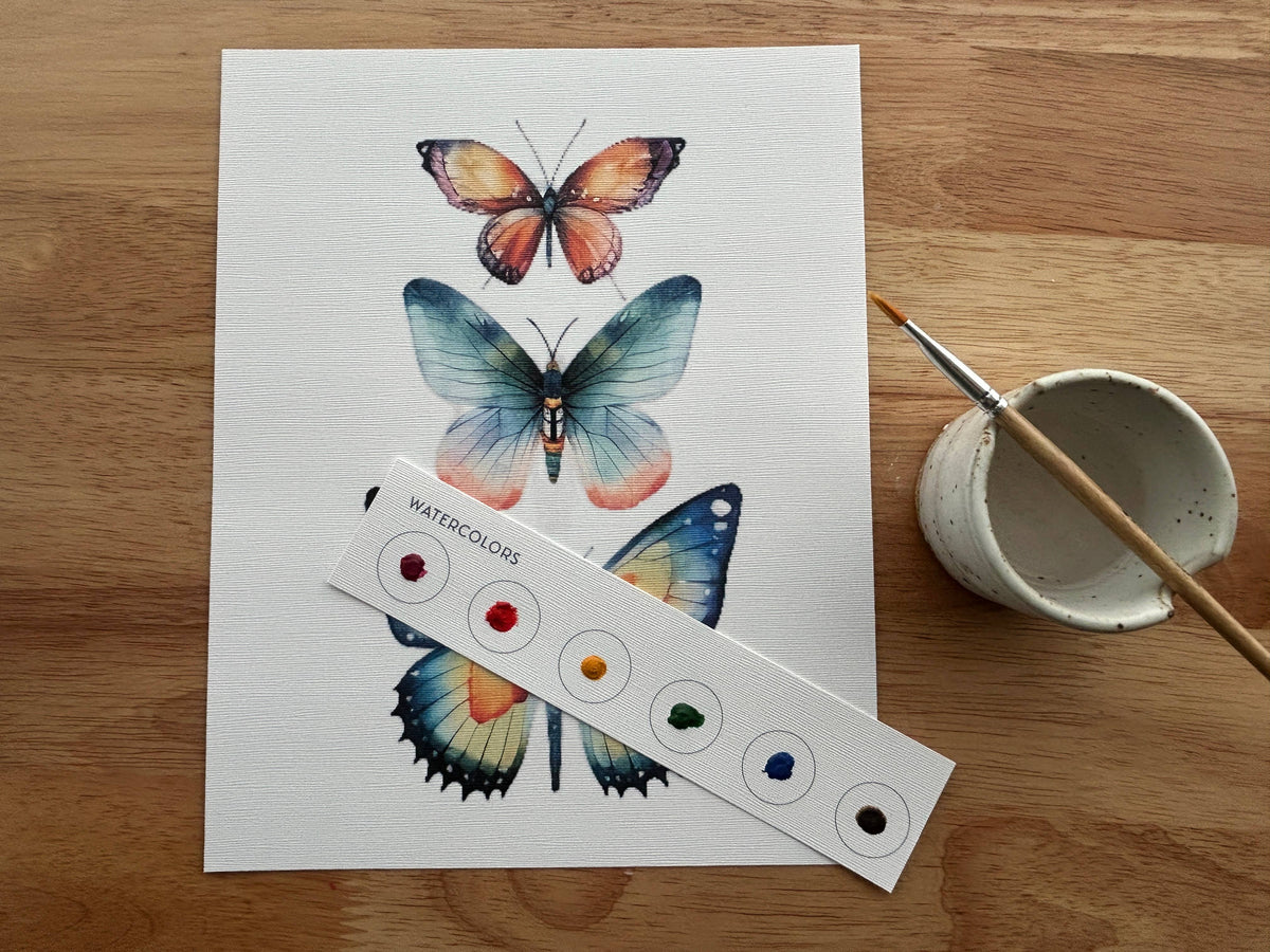 The Butterflies All-In-One Beginner Watercolor Kit by Breathe People is shown with a painting of three colorful butterflies on a wooden table, beside a paintbrush and small water cup.