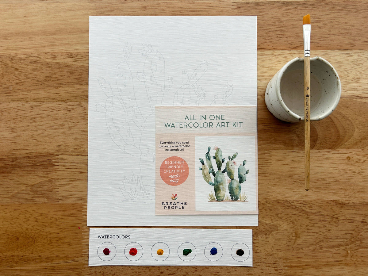 The Breathe People Desert Cactus All-In-One Beginner Watercolor Kit includes a paintbrush in a cup, cactus outline sheet, five watercolor paints, and a card with a painted cactus—perfect for easy watercolor practice on any surface.