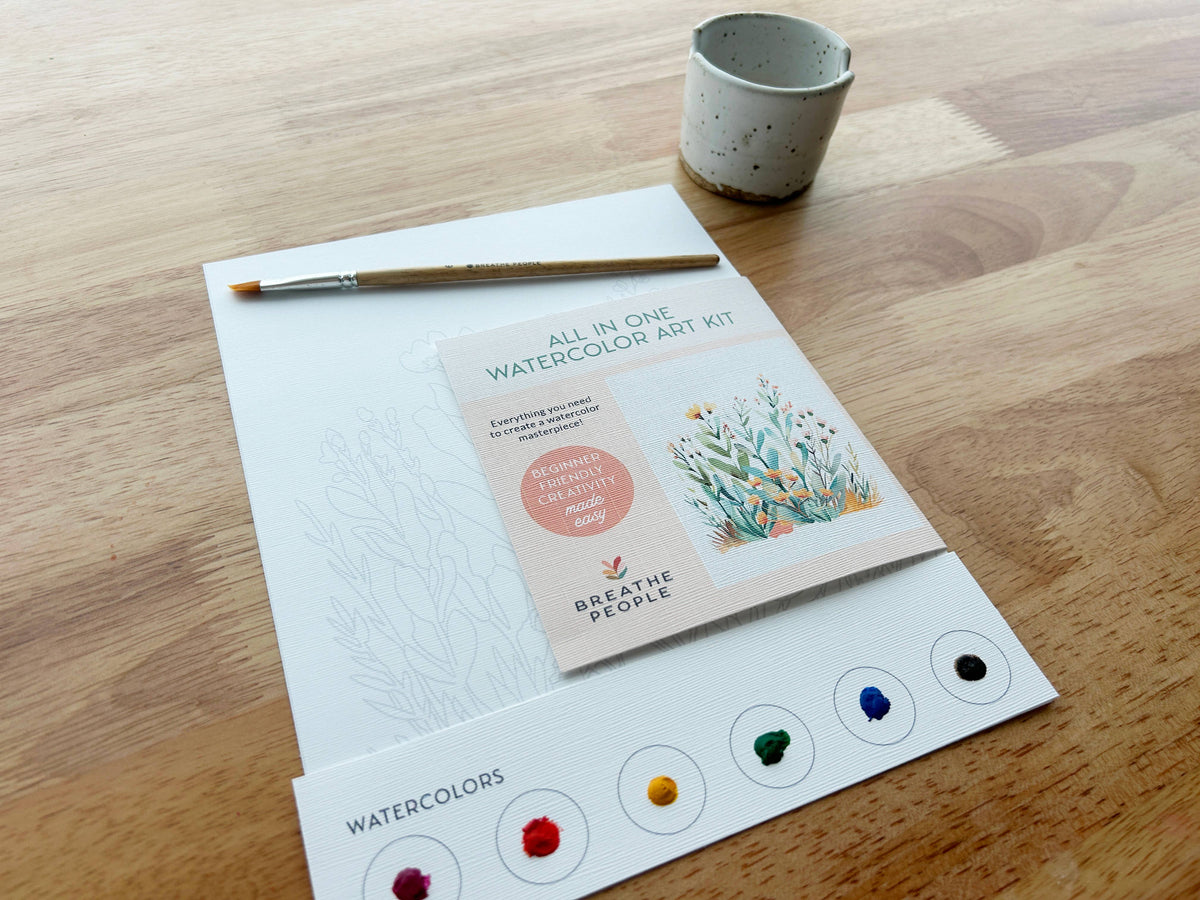 The Breathe People Desert Cactus All-In-One Beginner Watercolor Kit sits on a wooden table with a paintbrush, ceramic water cup, floral outline sheet, practice guide, and six vibrant paint dots—everything you need to create your masterpiece.