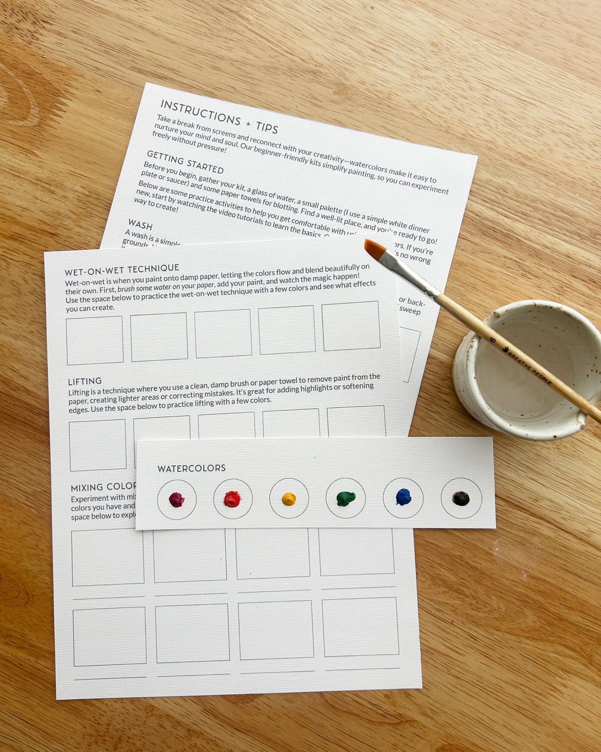The Breathe People Desert Cactus All-In-One Beginner Watercolor Kit includes a painting setup with instructions, a paintbrush, water bowl, and a card with five watercolor paint dots (yellow, red, pink, blue, green) on wood.