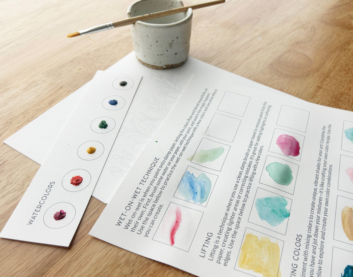 A close-up of the Breathe People All-In-One Beginner Watercolor Kit - Hummingbird on a wooden surface, showing a paintbrush on a ceramic cup, paint sample cards, and instructional sheets with watercolor techniques.