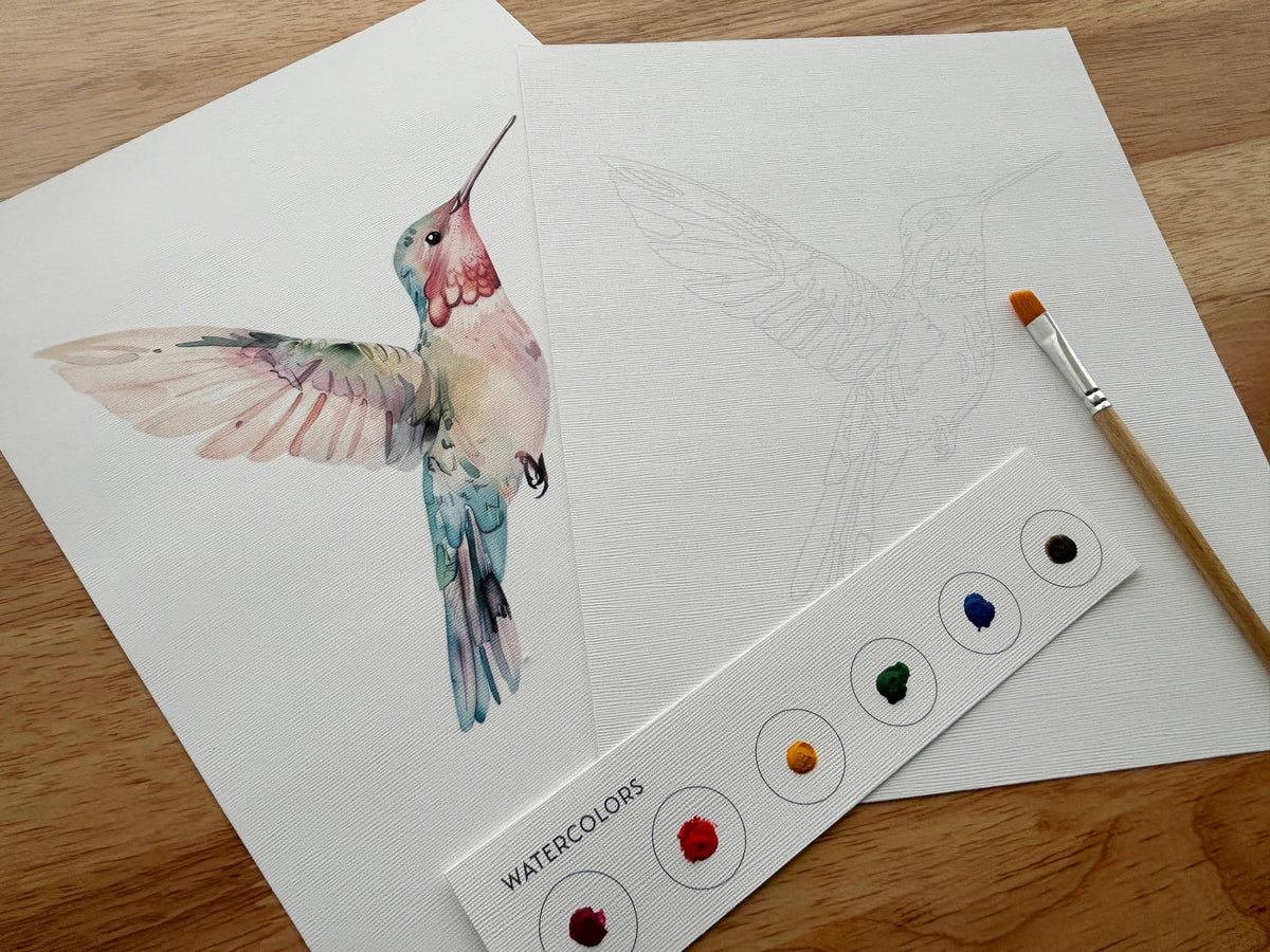On a wooden surface, two sheets from the Breathe People All-In-One Beginner Watercolor Kit - Hummingbird show a vibrant painting and pencil sketch, with a paintbrush and included watercolor paints beside them.