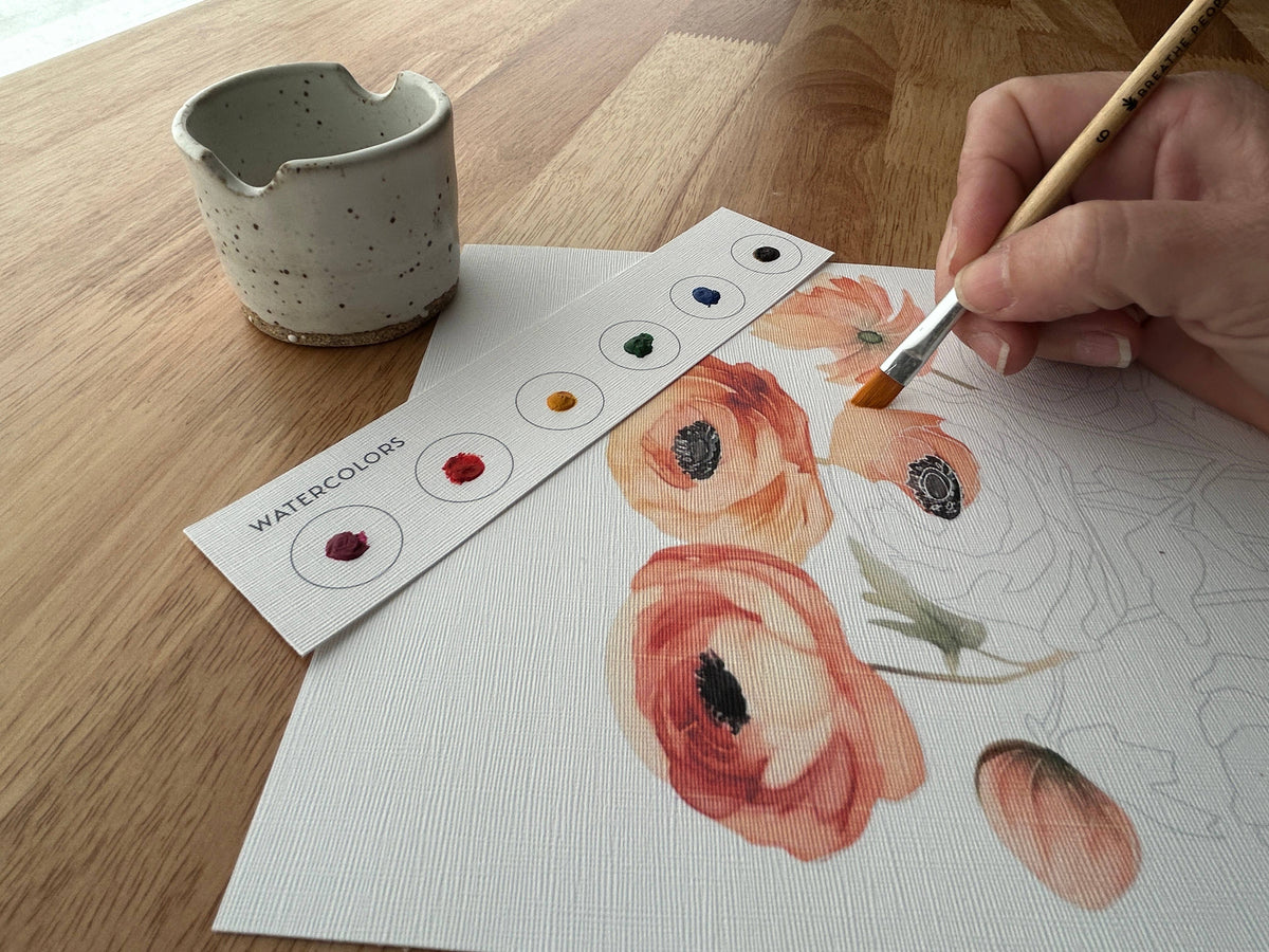 A hand uses a paintbrush from Breathe Peoples Mushroom Forest All-In-One Watercolor Kit to paint orange and pink flowers on textured paper, with a palette card and ceramic cup on a wooden table.