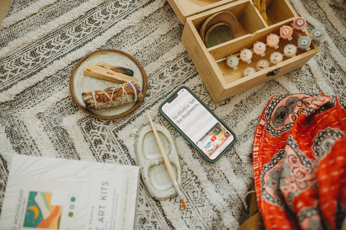 A phone with a mindfulness app rests on a patterned rug beside Breathe People’s Night Mushrooms Paint by Number Kit, Palo Santo sticks, a paintbrush, the kit box, and a red scarf, creating a cozy creative vibe.