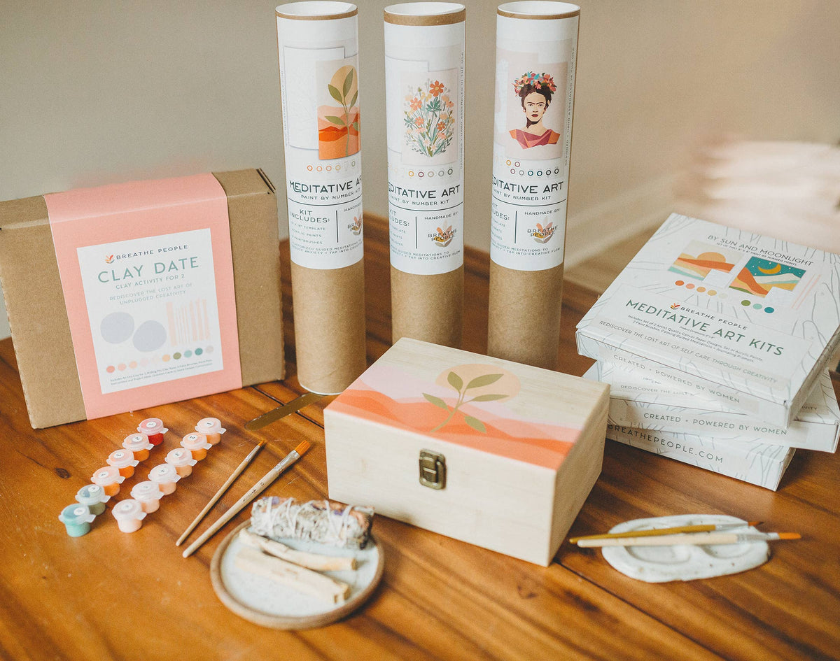 A meditative art kit collection on a wooden table, featuring Breathe Peoples Night Mushrooms Paint by Number Kit, a “Clay Date” kit, plant-etched wooden box, paintbrushes, paint pots, tools, and illustrated instructions.
