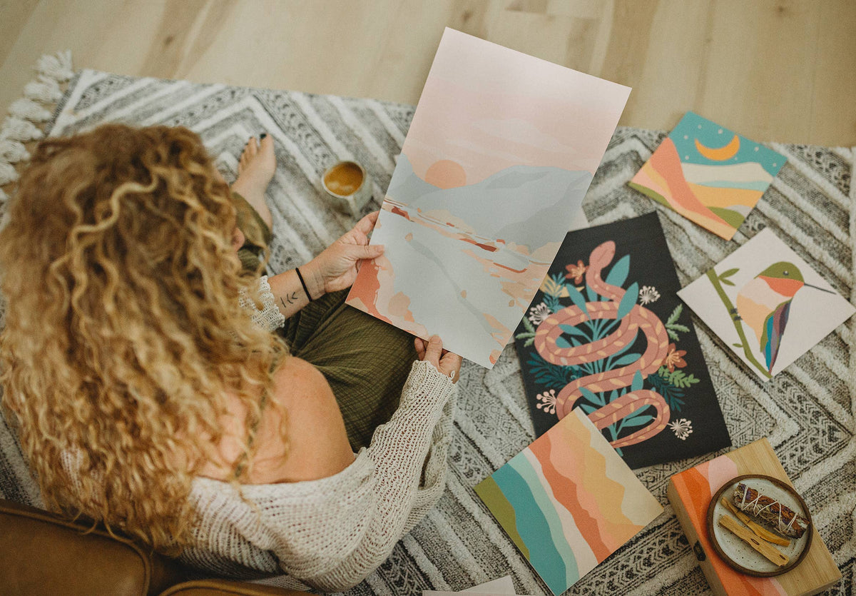 A person with curly hair sits on the floor, holding the Night Mushrooms Paint by Number Kit from Breathe People. Colorful artworks—including meditative art, a snake, bird, and abstracts—are spread out on the patterned rug around them.