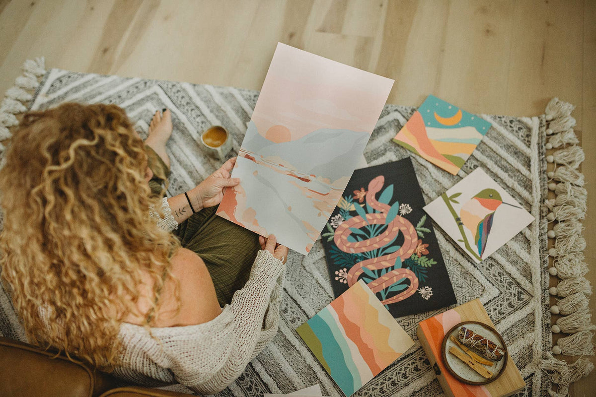 A person with curly hair sits on a patterned rug, holding the Vibrant Butterflies Paint by Number Kit from Breathe People. Around them are colorful meditative and nature-inspired artworks, plus a cup of tea.