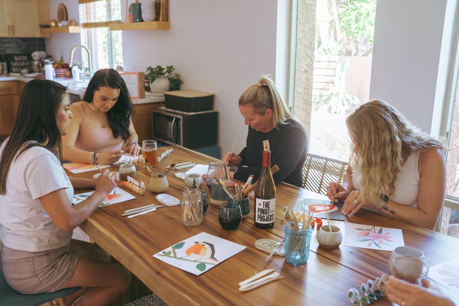 Four women sit around a wooden table in a bright kitchen, enjoying the Vibrant Butterflies Paint by Number Kit from Breathe People. Art supplies, drinks, and completed butterfly paintings are scattered across the table.