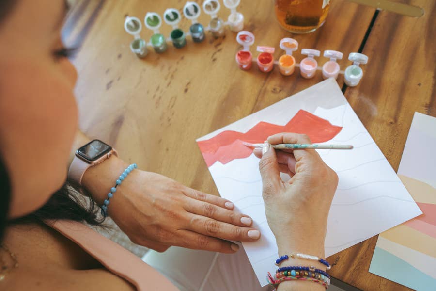 Wearing bracelets and a smartwatch, a person relaxes by painting the Vibrant Butterflies Paint by Number Kit from Breathe People at a wooden table surrounded by open paint pots.