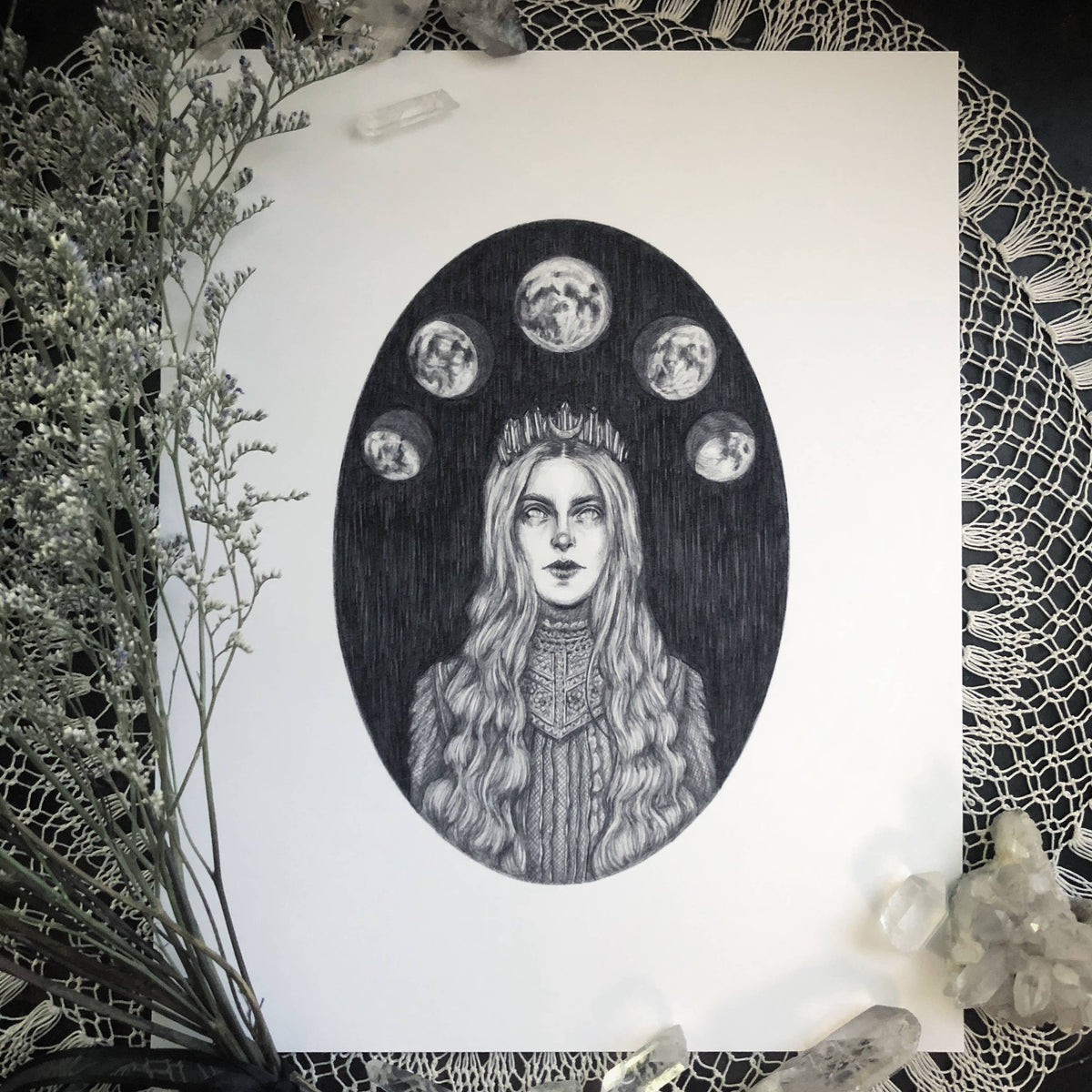 Selene Fine Art Print (10x8) by Caitlin McCarthy Art features a mystical long-haired woman in a crown, inspired by the Greek Moon Goddess. Five lunar phases, lace, crystals, and dried foliage frame this black-and-white work on archival matte paper.