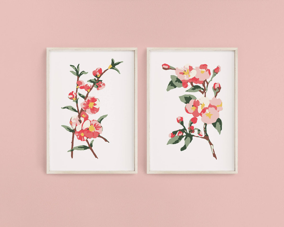 Display two framed prints created with the Cate Paper Co. DIY Art Cherry Blossoms Paint-By-Numbers Kit, featuring pink and red flowers with green leaves on a pale pink wall in a minimalist, paint-by-number style.