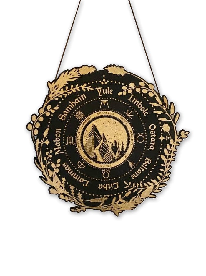 The Wheel of the Year, Pagan Calendar: Stained Black by CSAcreative is a round birch wood wall plaque with gold designs on black. Sabbat names surround a geometric mountain and moon motif, making it a striking decor piece to hang.
