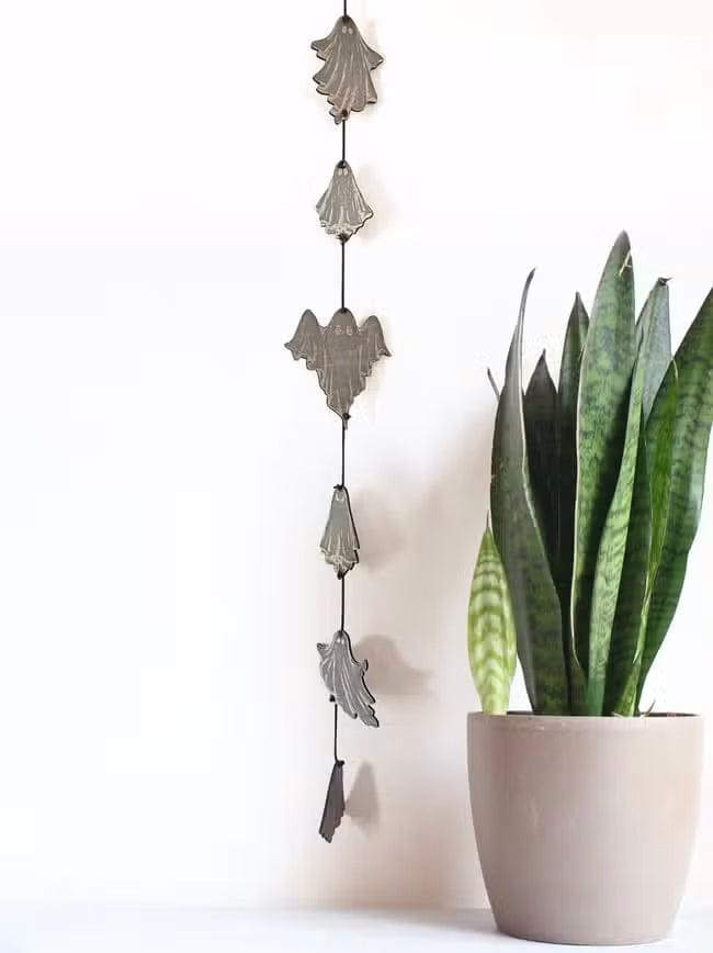 The CSAcreative Ghost Garland, featuring metal leaf, bat, and engraved ghost ornaments, hangs on a white wall beside a snake plant in a beige pot. The minimalistic and bright setup is ideal for subtle Halloween decor.
