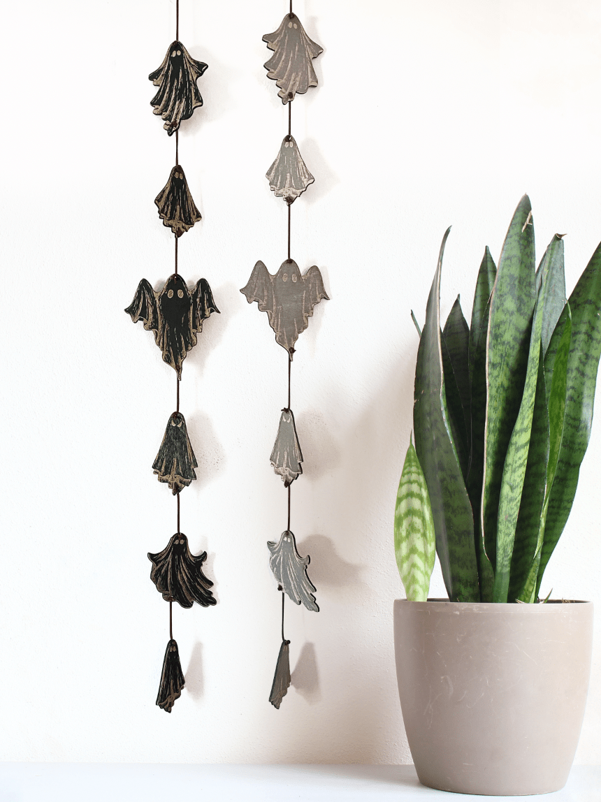 A potted snake plant sits beside two CSAcreative Ghost Garlands featuring engraved ghost and bat shapes, creating an ideal Halloween display against a white wall.