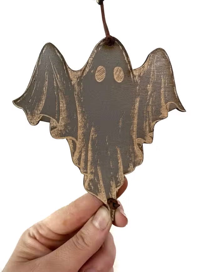 A hand holds a CSAcreative Ghost Garland wooden ornament, shaped like a ghost with rounded wings and two circular eyes, perfect for Halloween decor, hanging by a brown string against a white background.