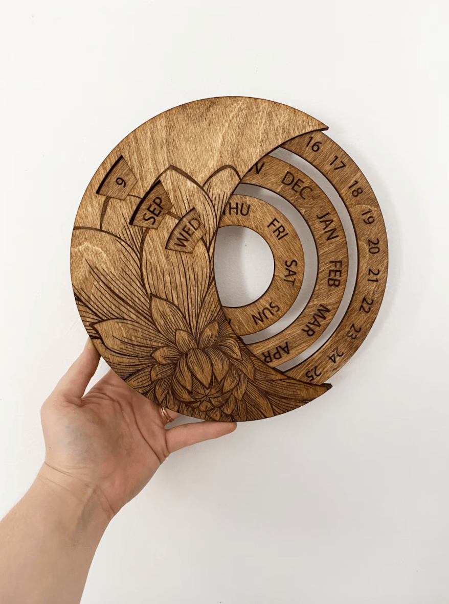 A hand holds a CSAcreative Perpetual Calendar, a sustainable wooden calendar with a floral design. Handcrafted, it features rotating rings to display days, months, and dates against a plain white background.