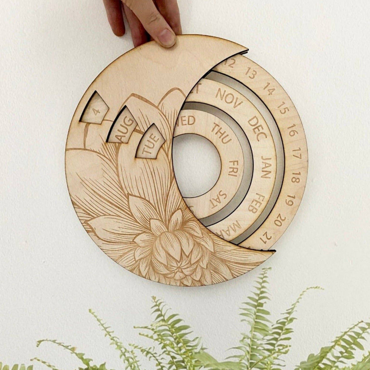 A hand holds the CSAcreative Perpetual Calendar, a sustainable wooden piece with rotating month and date rings, a triangular marker, and intricate floral carvings. Ferns appear at the bottom of the image.