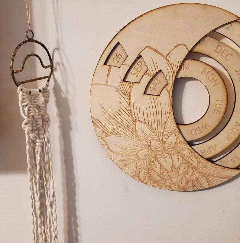 The CSAcreative Perpetual Calendar, a wooden piece with engraved florals showing 26 Sep Sat, sits beside a decorative macramé with metal top against a light wall.