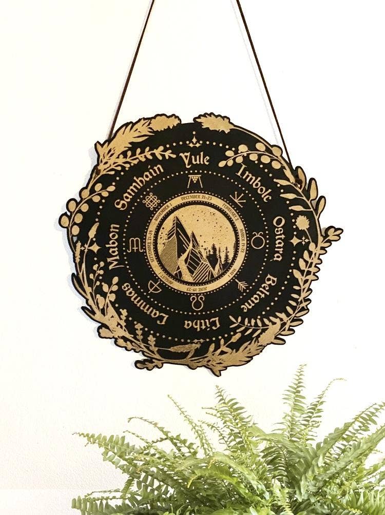 The Wheel of the Year, Pagan Calendar: Stained Black wall art by CSAcreative features pagan sabbats and zodiac symbols encircling a mountain on birch wood, with a green fern below on white.