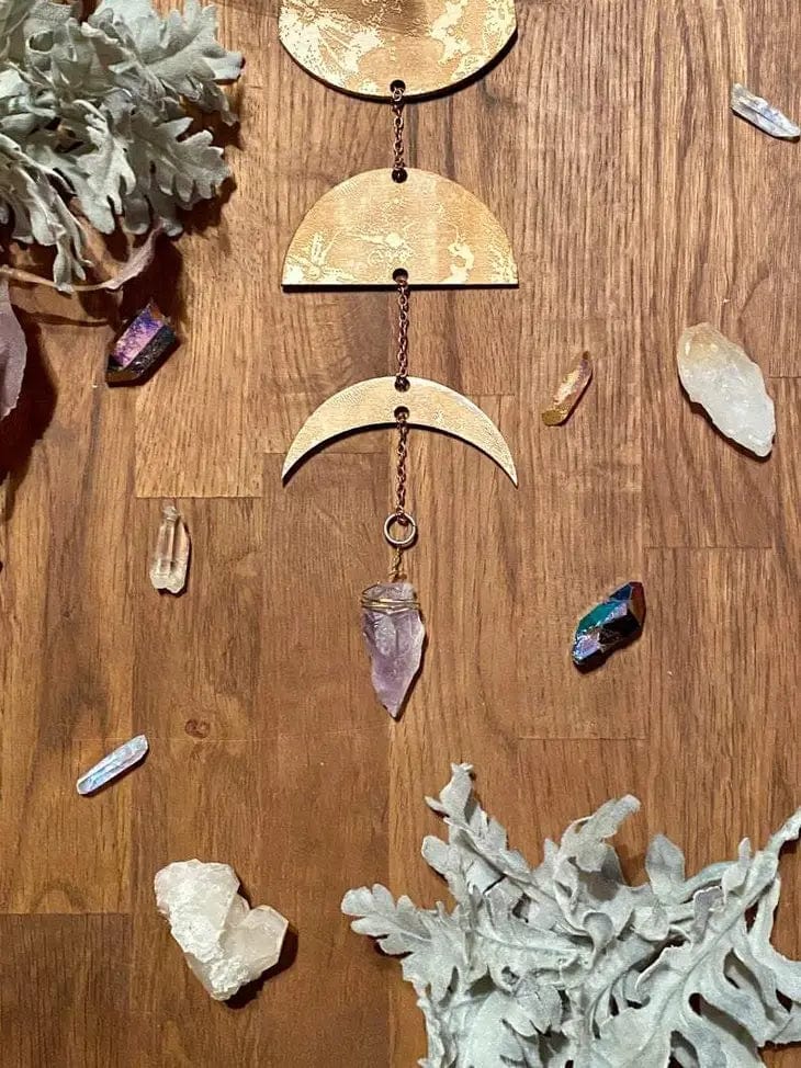 A birch wood surface features CSAcreatives Gold Floral Phases of the Moon Garland with Amethyst, accented by clear and iridescent crystals and two clusters of pale, silvery leaves.