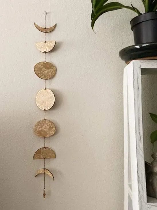The CSAcreative Gold Floral Phases of the Moon Garland with Amethyst hangs on a beige wall near a white shelf and a potted plant.